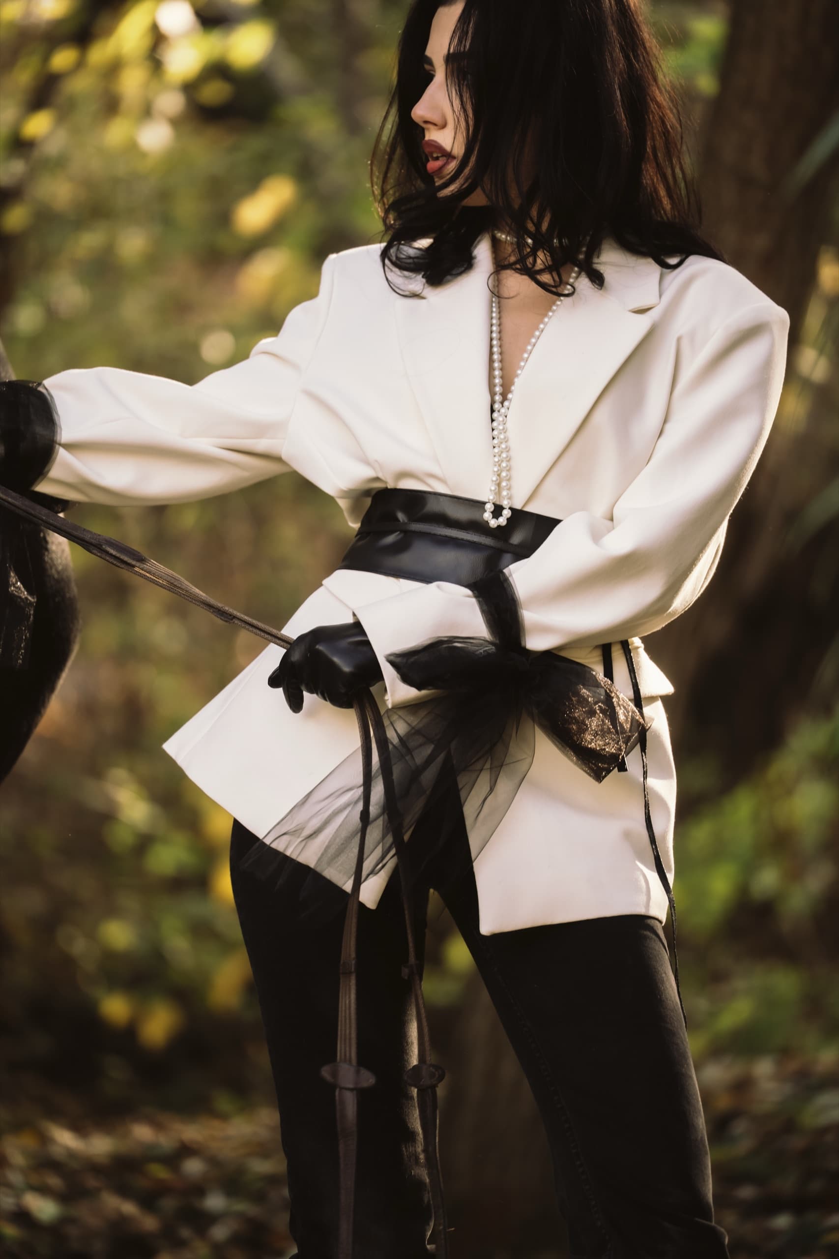 Cream blazer cinched with a wide black belt and tulle bow, beside a horse in autumn woods