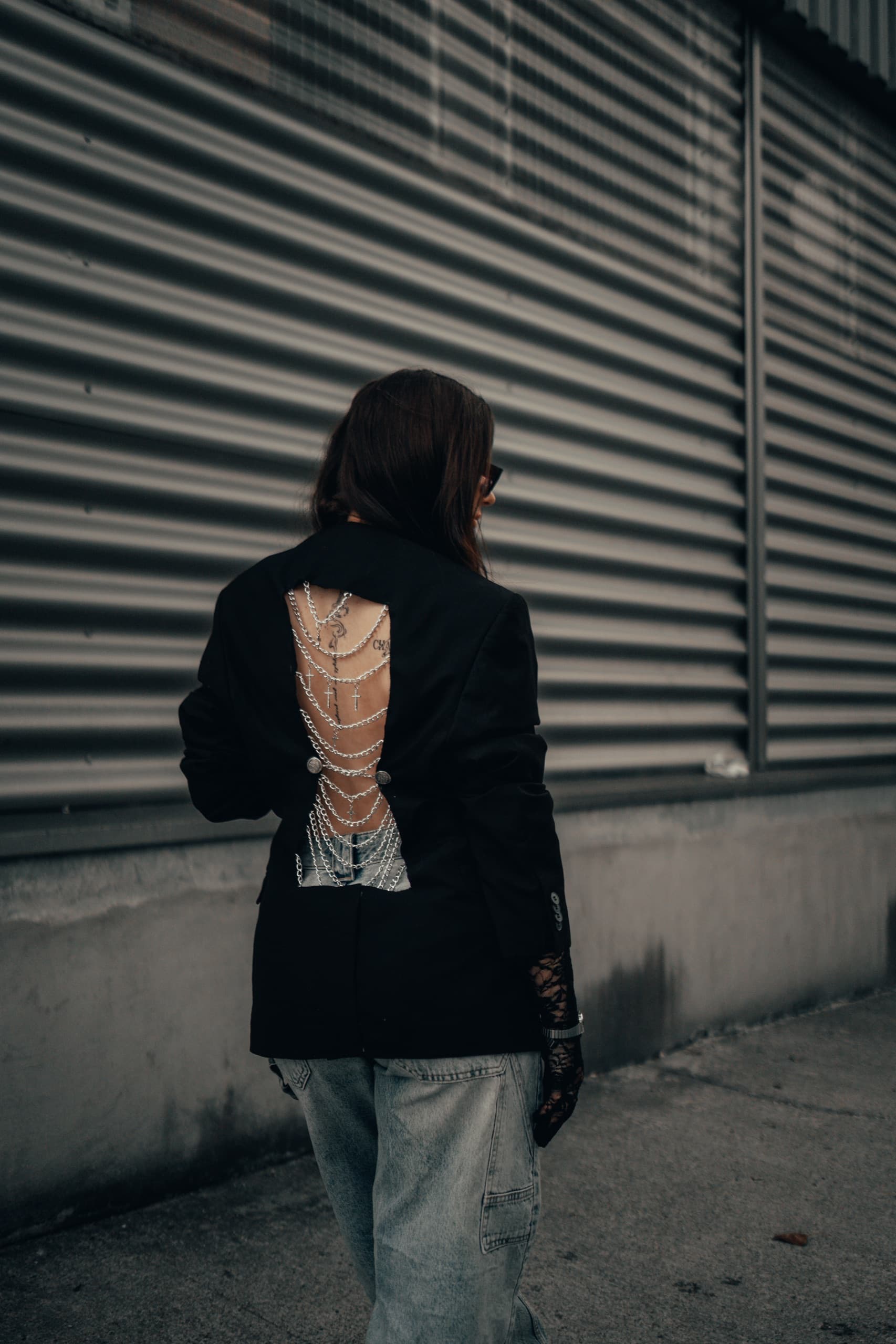 Back view of a black blazer with an open back laced with layered chains and cross charms, against corrugated metal