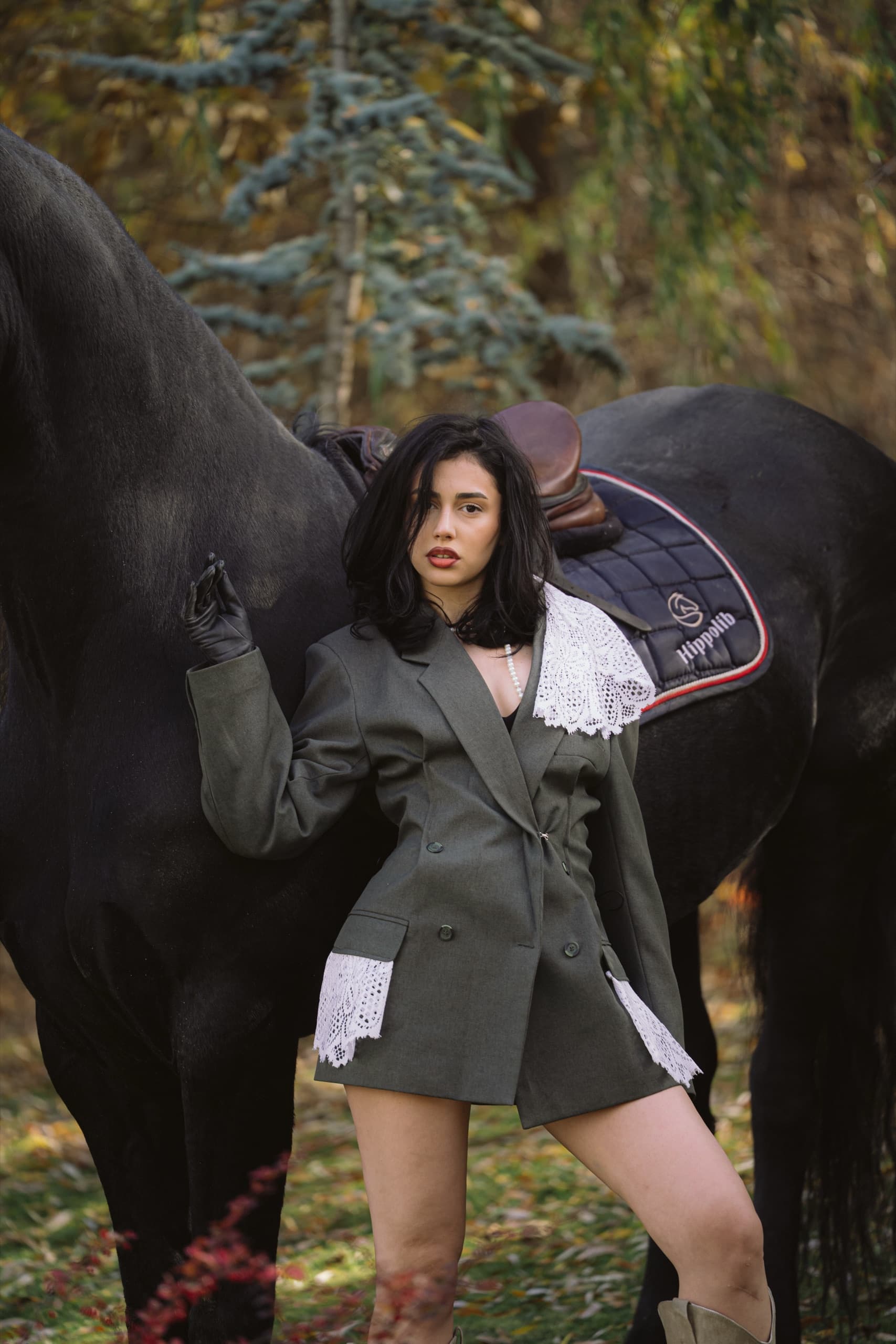Olive double-breasted blazer with white crochet lace details, in front of a black horse in autumn woods