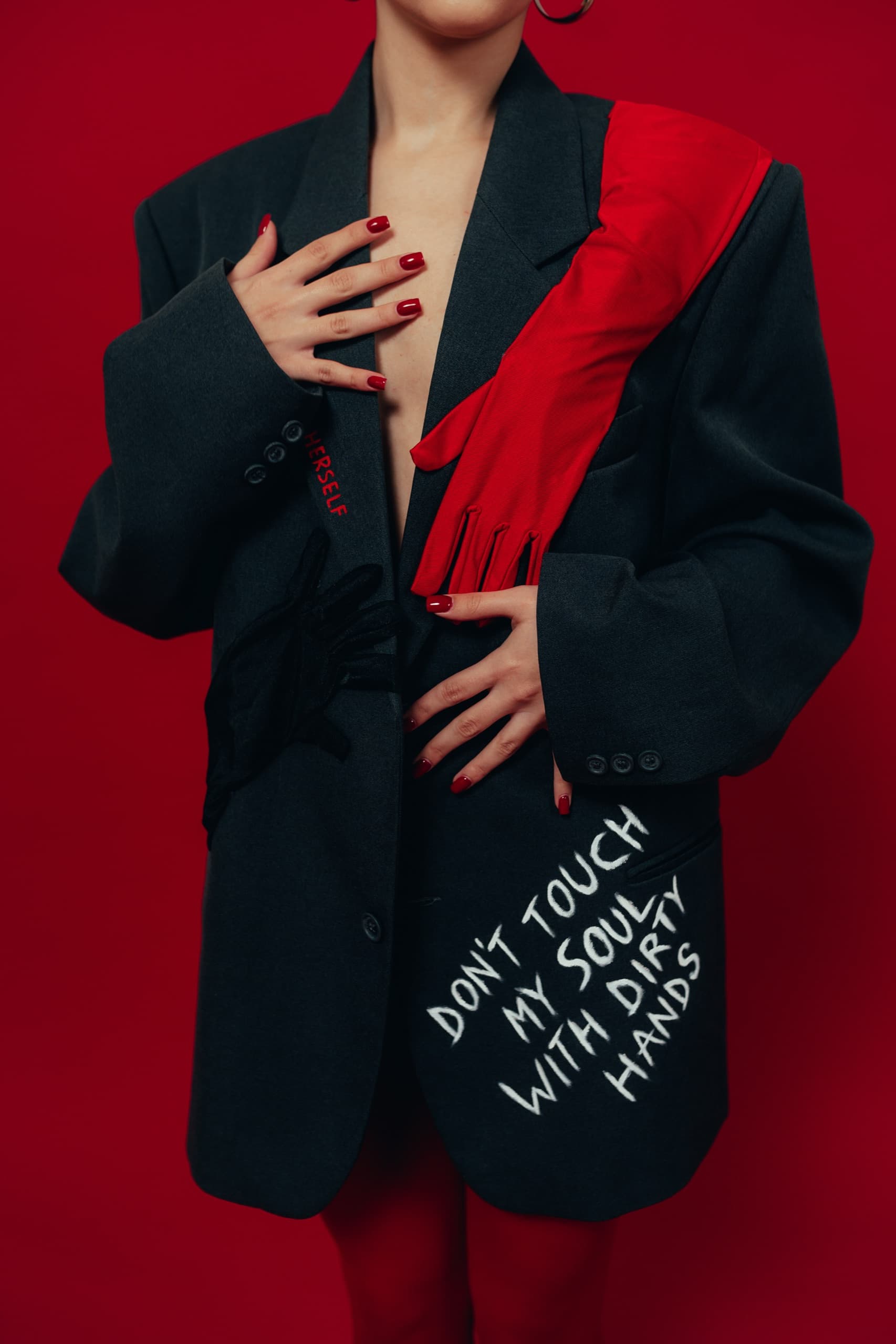 Detail of a charcoal blazer with glove appliques and a hand-painted slogan on the hem, against a deep red backdrop