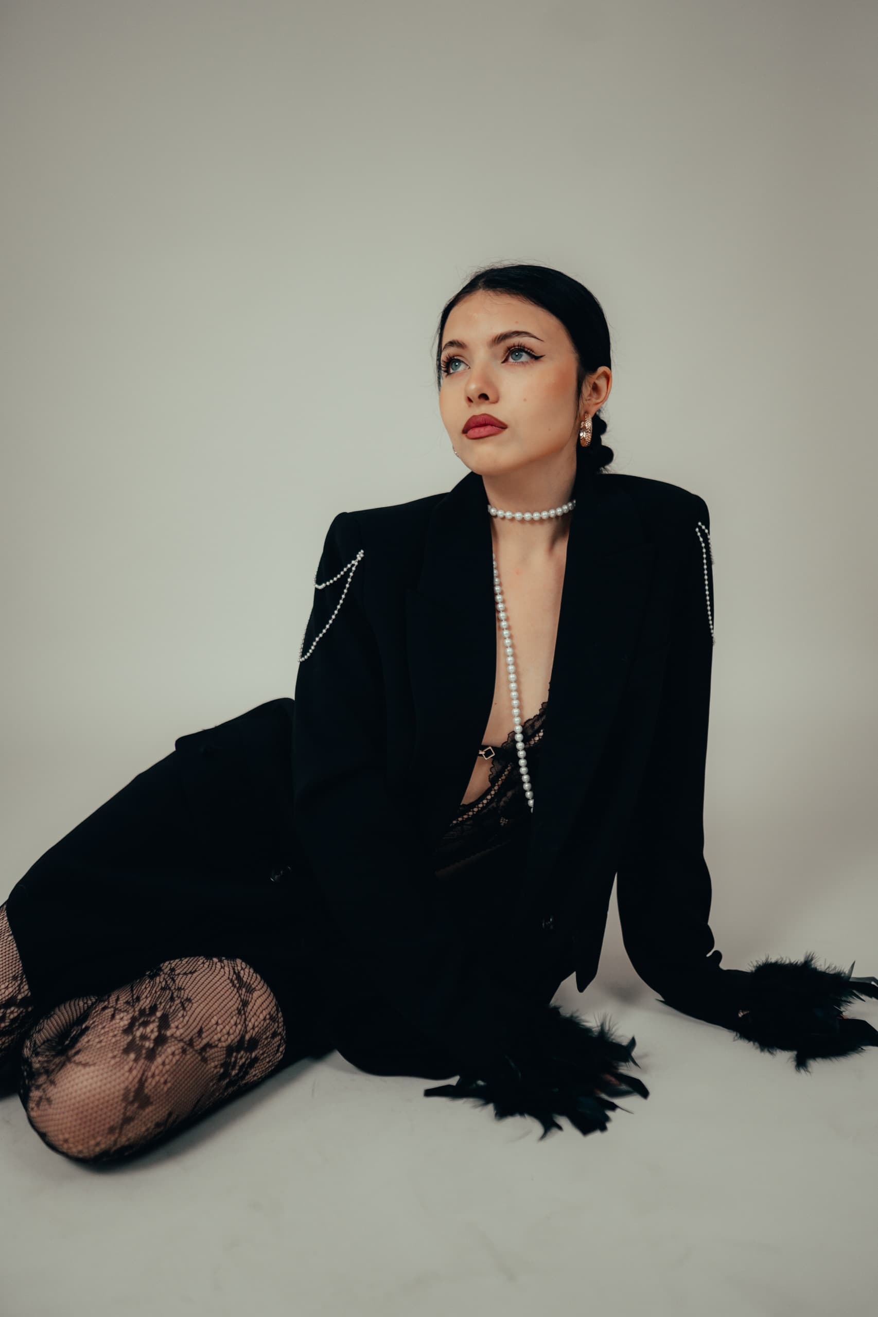 Black blazer with draped pearl chains and feather-trimmed cuffs, model seated on a studio floor