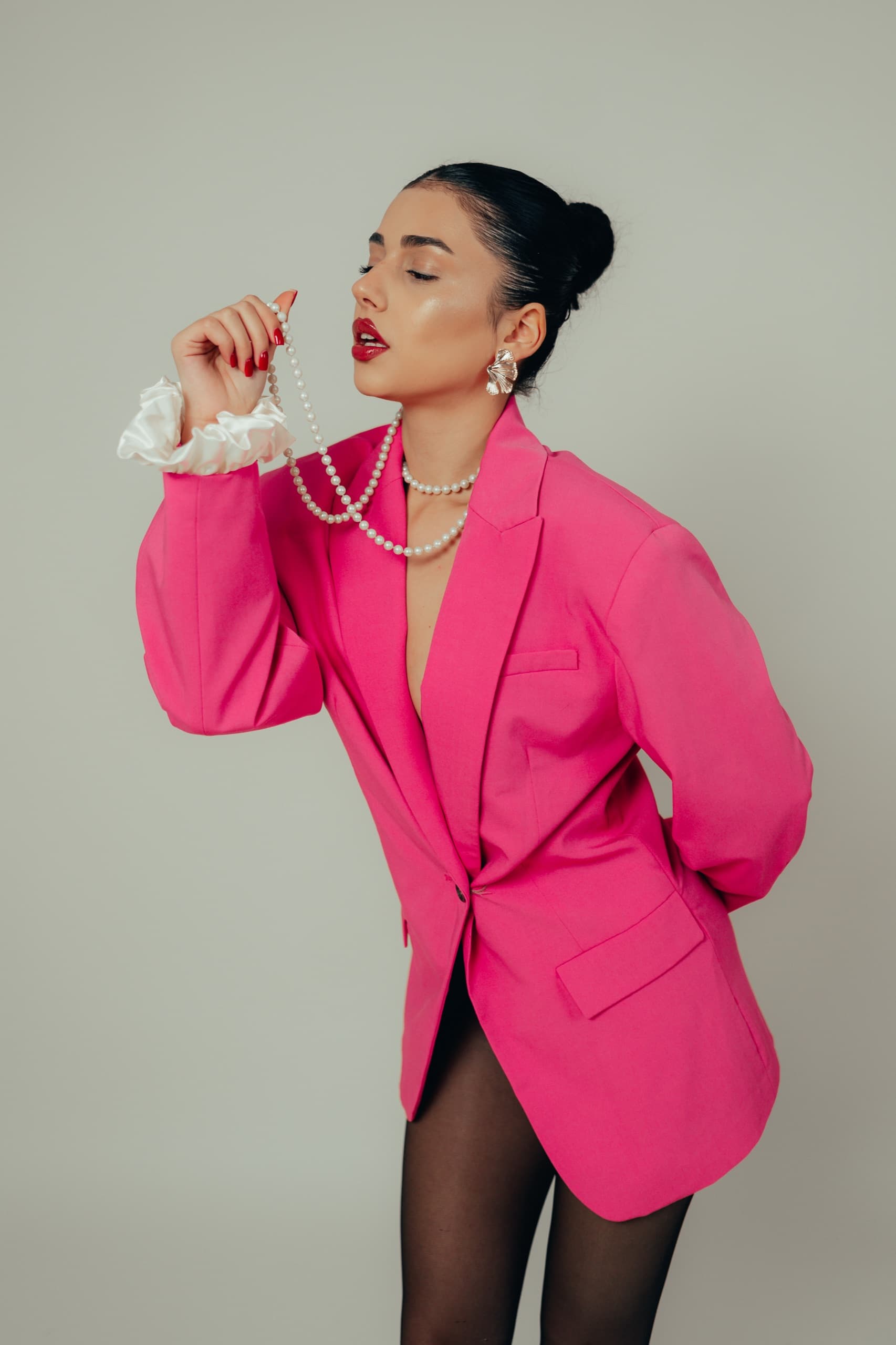 Hot pink blazer with a white satin ruffle cuff, model lifting a long pearl strand