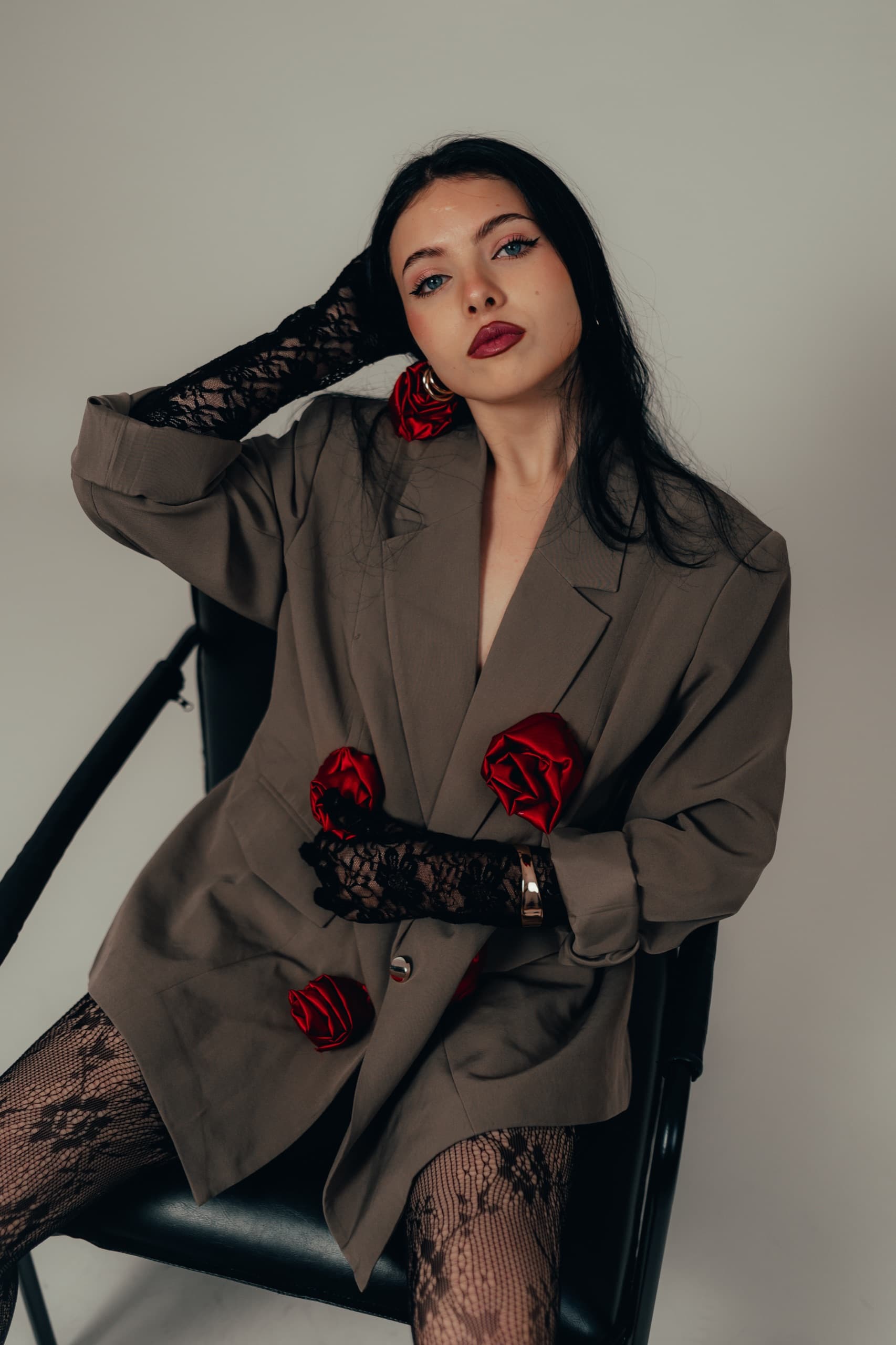 Taupe blazer with red satin rose appliques, model reclining on a black chair with lace gloves