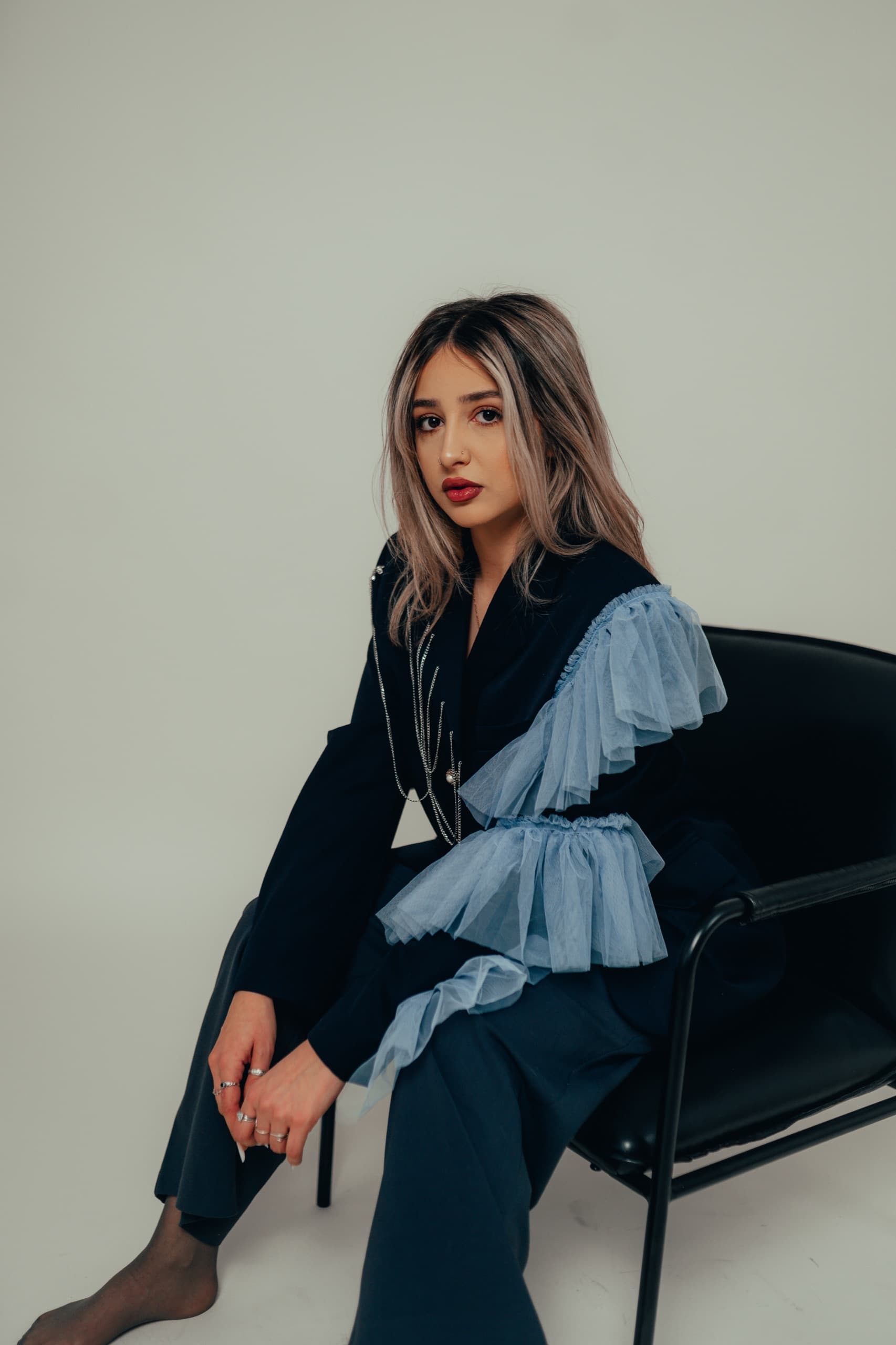 Navy blazer with tiered light blue tulle ruffles and a chain lapel detail, model seated on a black chair