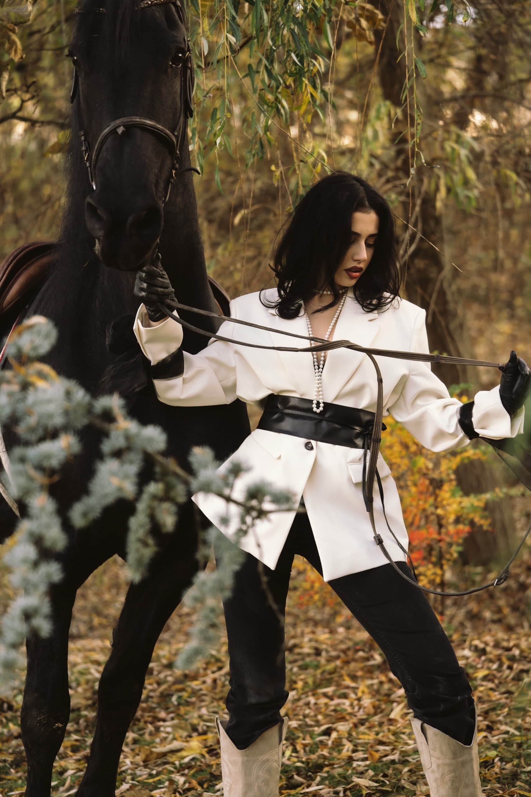 A white blazer belted in black leather, beside a black horse in autumn woods