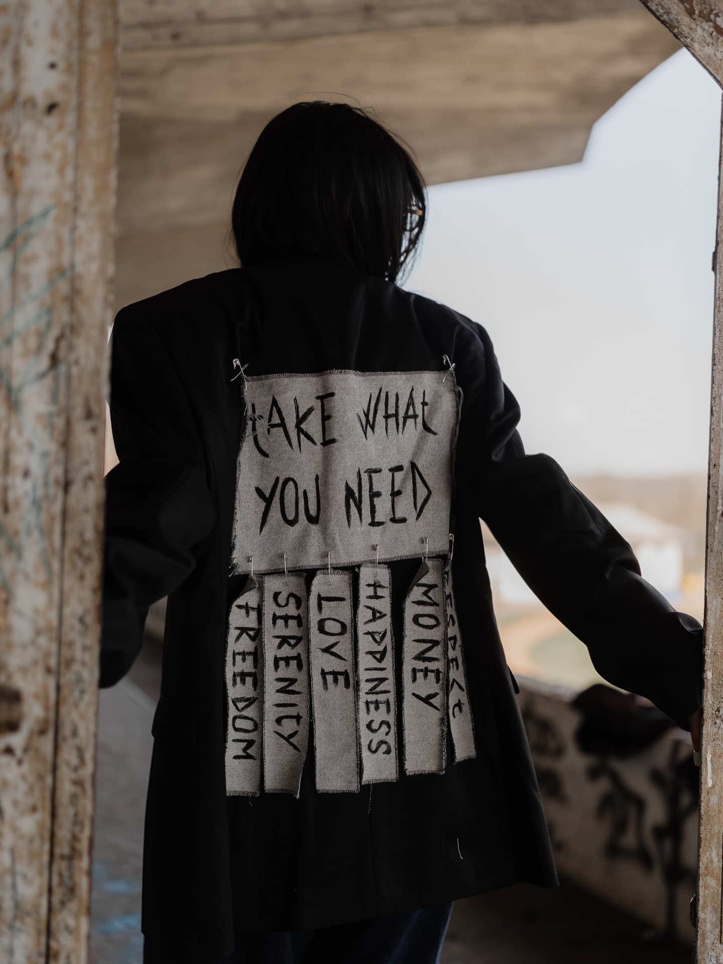 Back of a black blazer with a hand-painted canvas patch reading take what you need, fixed with safety pins