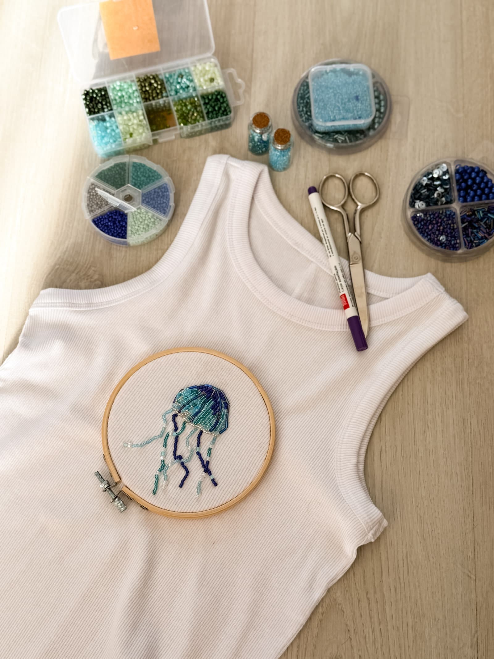 A white ribbed tank on a work table with a beaded jellyfish half stitched in an embroidery hoop, beside bead trays, scissors and a marker