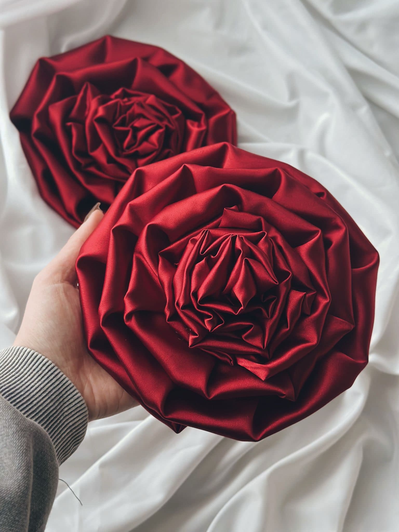 Hands holding two large brooches folded from deep red satin into roses, on white cloth
