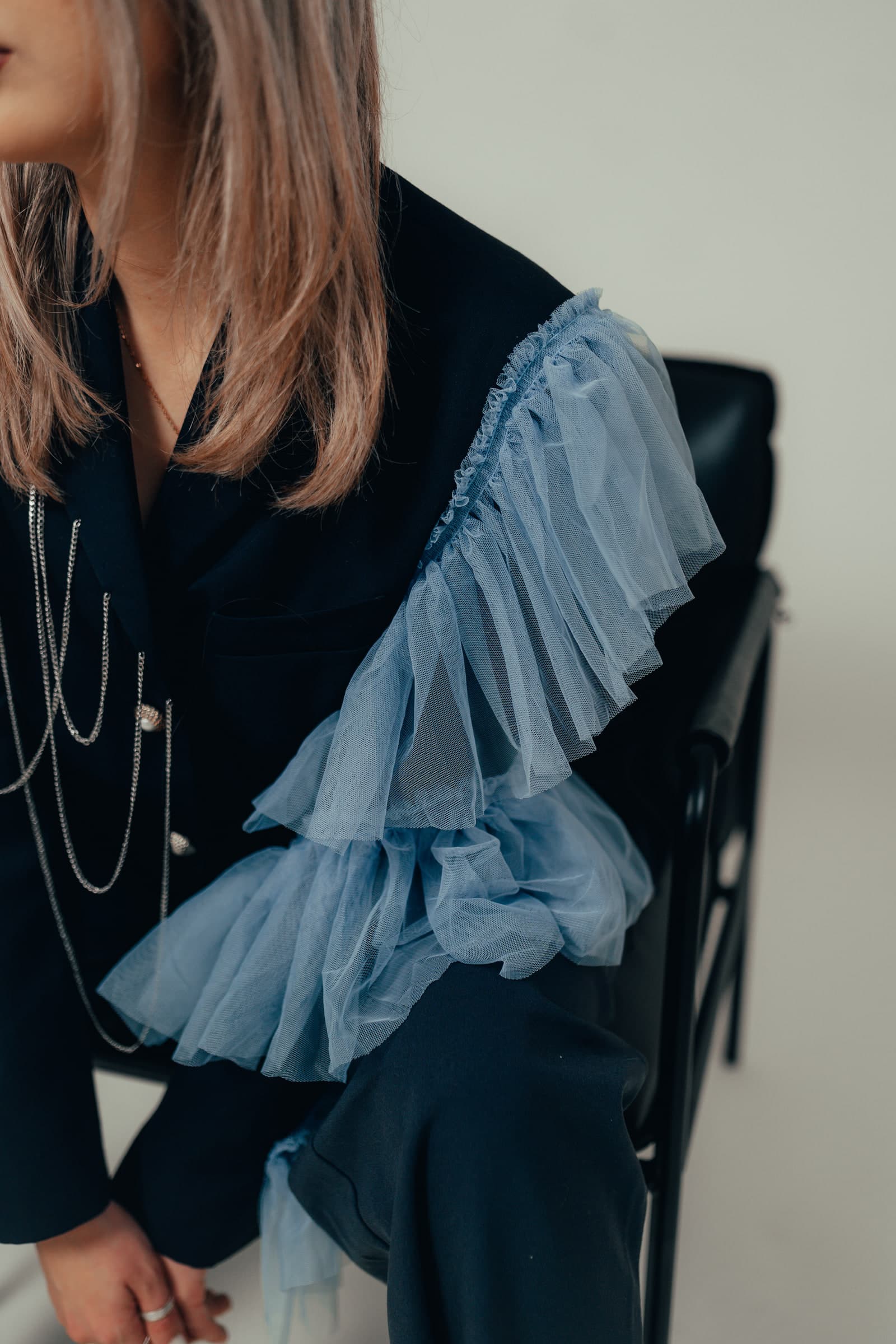 A black jacket with an oversized powder blue tulle ruffle running down one sleeve, worn with layered pearl chains