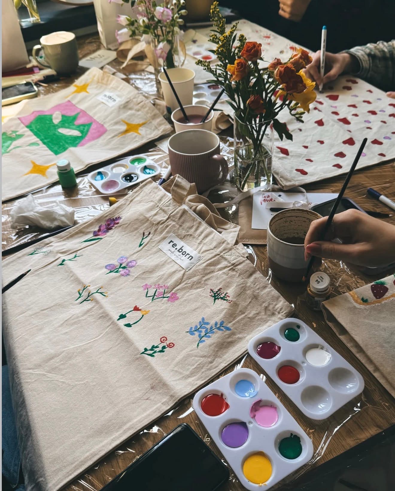 A workshop table laid with hand-painted tote bags, paints, brushes and flowers
